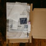 Open cardboard box with medical testing kit materials, including a security seal, packaging with biohazard symbol, and a capped vial, on a wooden table.