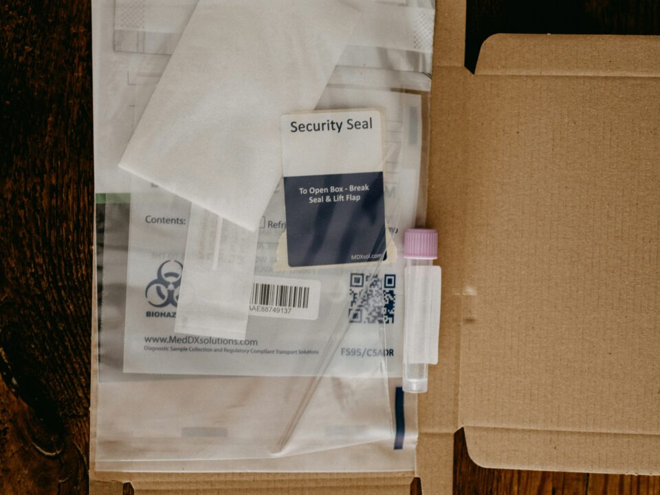 Open cardboard box with medical testing kit materials, including a security seal, packaging with biohazard symbol, and a capped vial, on a wooden table.