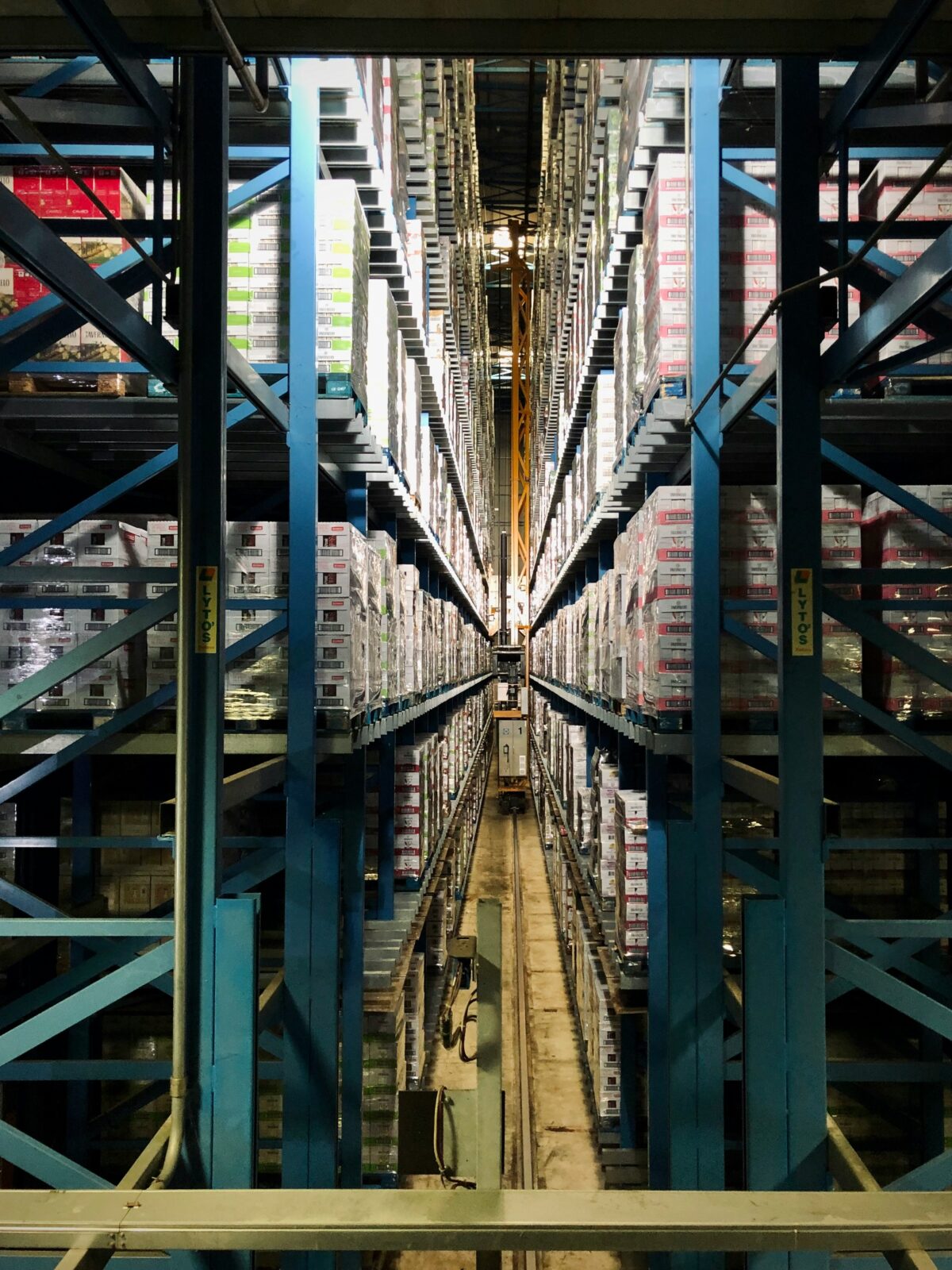 A vast warehouse with towering blue shelves filled with boxes. A narrow aisle runs down the center, showcasing a symmetrical, organized layout.