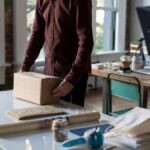 A person in a red shirt wraps a parcel in an office with a bright window. Nearby, there are papers, tape, and a laptop on a wooden table, creating a focused, busy atmosphere.