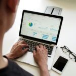 Person working on a laptop displaying data analytics with colorful charts and graphs. Nearby, glasses and a smartphone rest on a white desk.