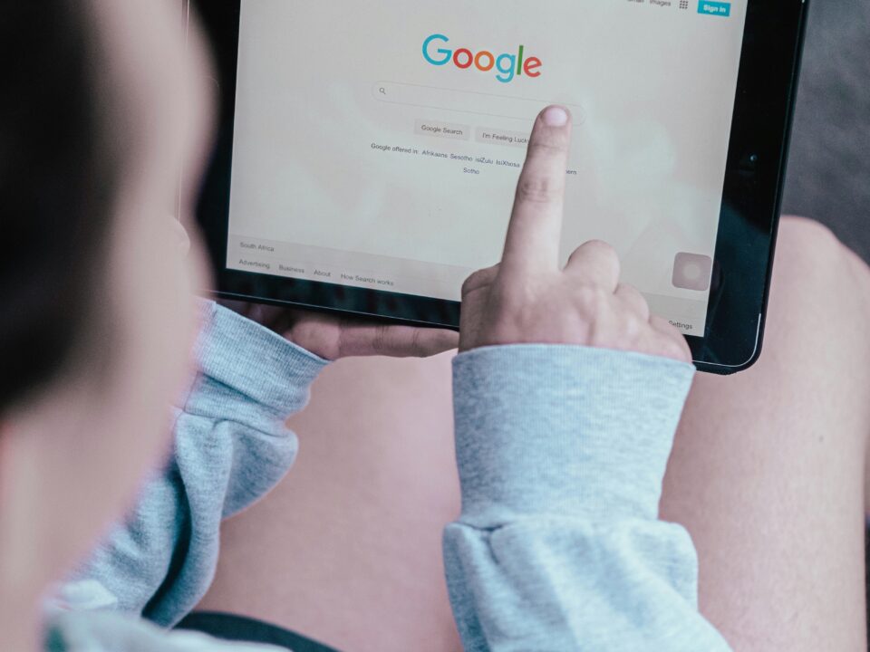A person in a grey sweatshirt uses a tablet, pointing at the Google homepage's search bar. The focus is on the screen, creating a tech-savvy vibe.