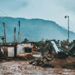 Industrial site with conveyor belts and metal structures under heavy rain, surrounded by muddy ground and mountains in the background.