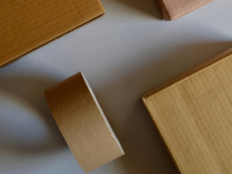Brown cardboard boxes and a roll of packing tape are arranged on a light gray surface. The scene conveys a sense of organization and preparedness.