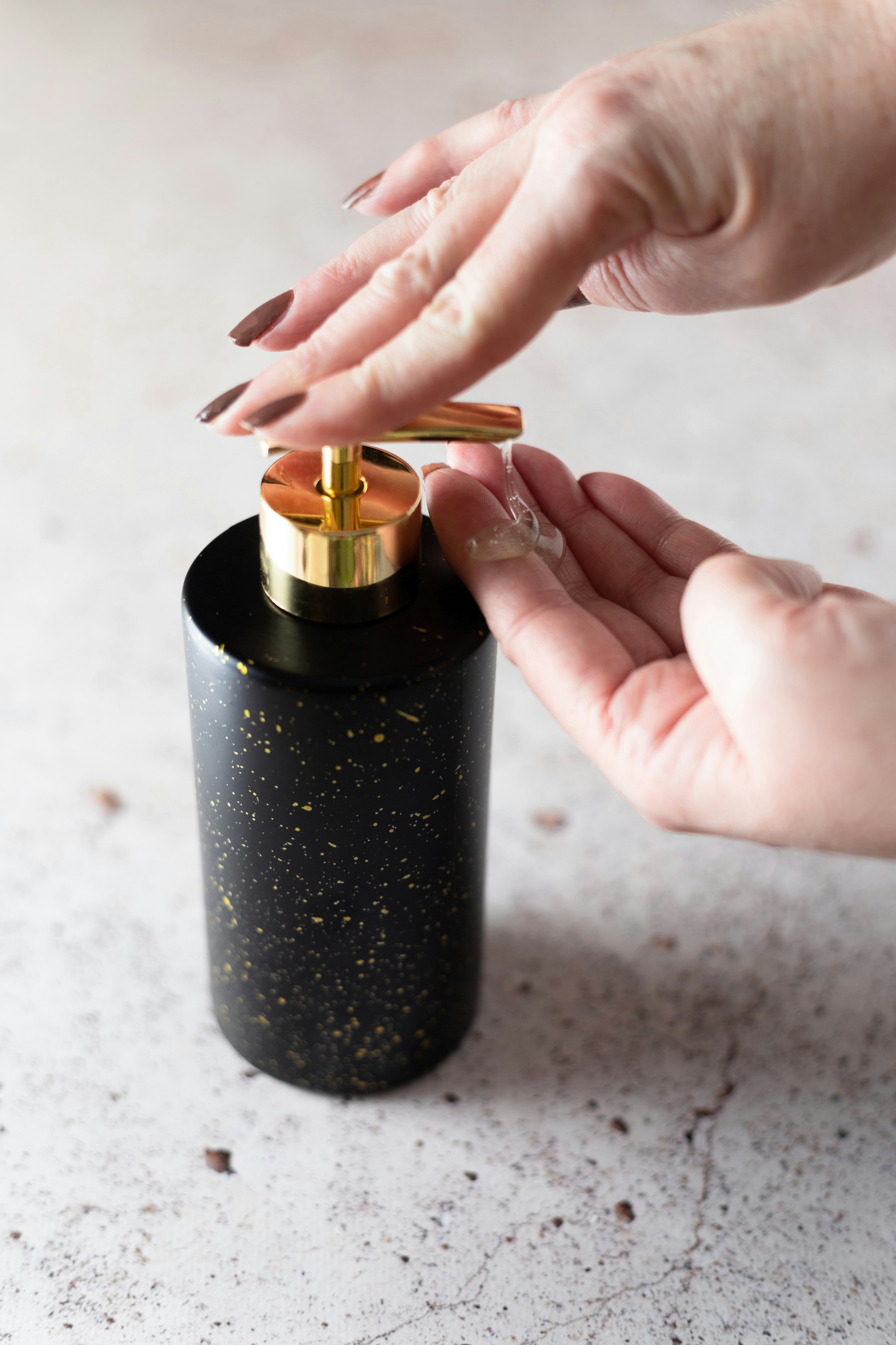Hands using a black and gold speckled soap dispenser on a light speckled countertop. The scene conveys cleanliness and elegance.