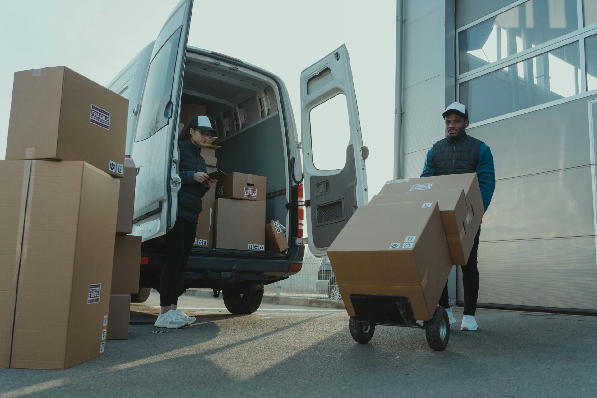 Two delivery workers are loading cardboard boxes labeled "fragile" into a van. One is inside the van, while the other uses a hand truck, conveying teamwork.