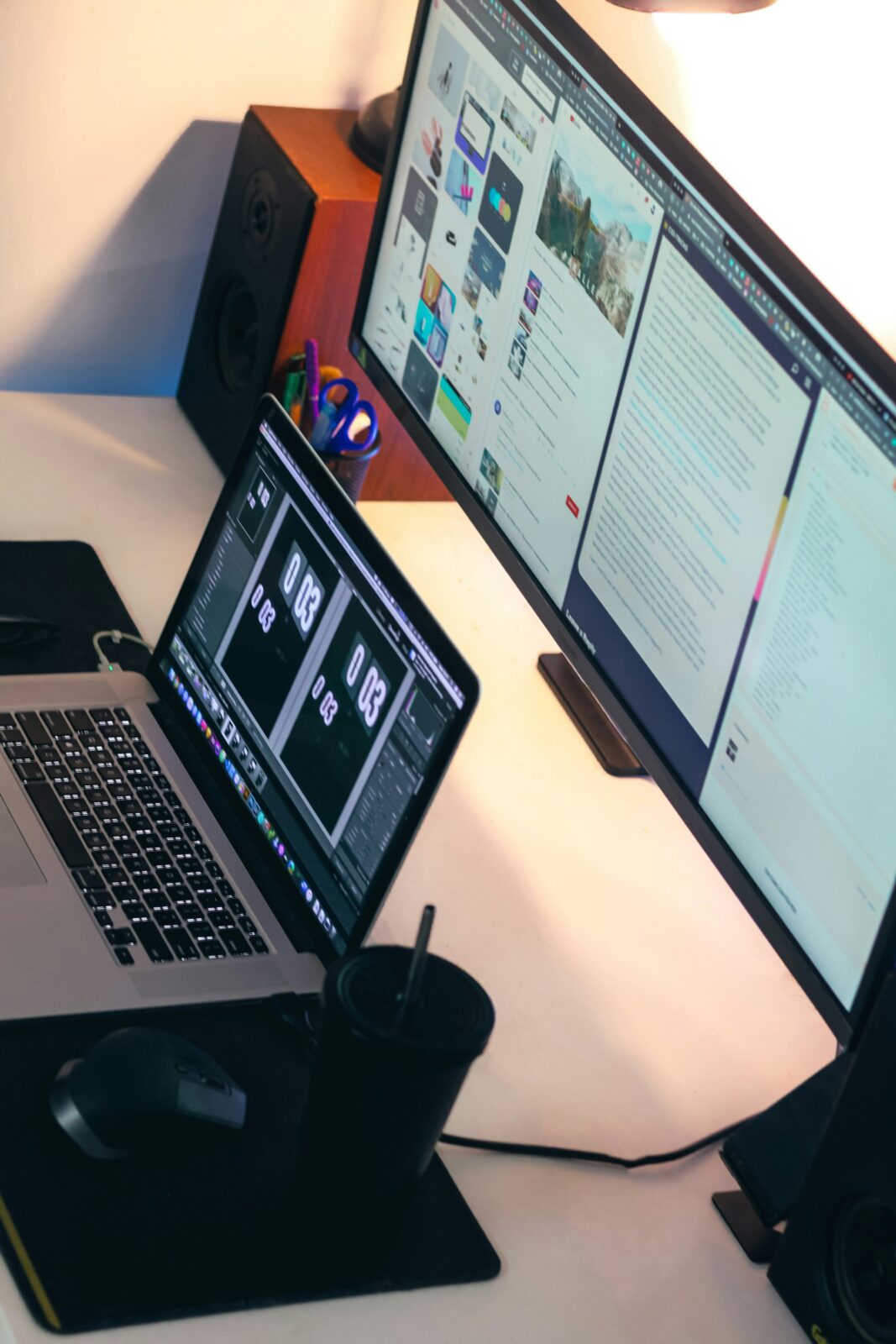 A workspace with a laptop and a large monitor displaying various digital content. A mouse, a black cup, and speakers are on the desk, conveying a tech-focused setup.