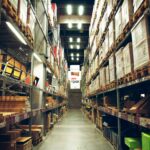 Wide warehouse aisle with towering shelves filled with boxed products. Bright overhead lights create an organized, industrial atmosphere.