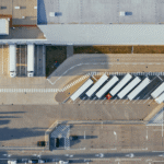 Aerial view of a modern European logistics hub with multiple trucks parked at loading docks of a large fulfillment warehouse.