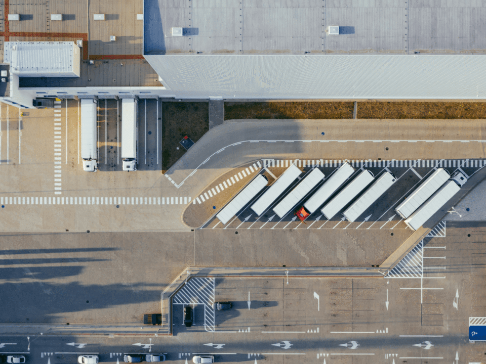 Aerial view of a modern European logistics hub with multiple trucks parked at loading docks of a large fulfillment warehouse.