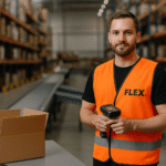 Warehouse worker wearing an orange safety vest with a black FLEX. logo stands in a modern fulfillment center, holding a barcode scanner in front of tall shelving racks filled with boxes.
