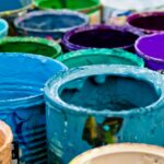 A close-up photograph of numerous open paint cans clustered together, their rims thickly coated with various colors including cyan, blue, purple, and green.