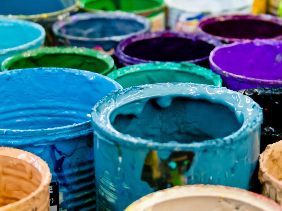 A close-up photograph of numerous open paint cans clustered together, their rims thickly coated with various colors including cyan, blue, purple, and green.
