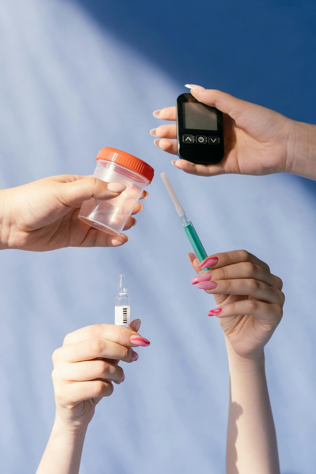 Four hands holding medical items: a glucometer, a specimen cup, an insulin vial, and a syringe, against a blue background.