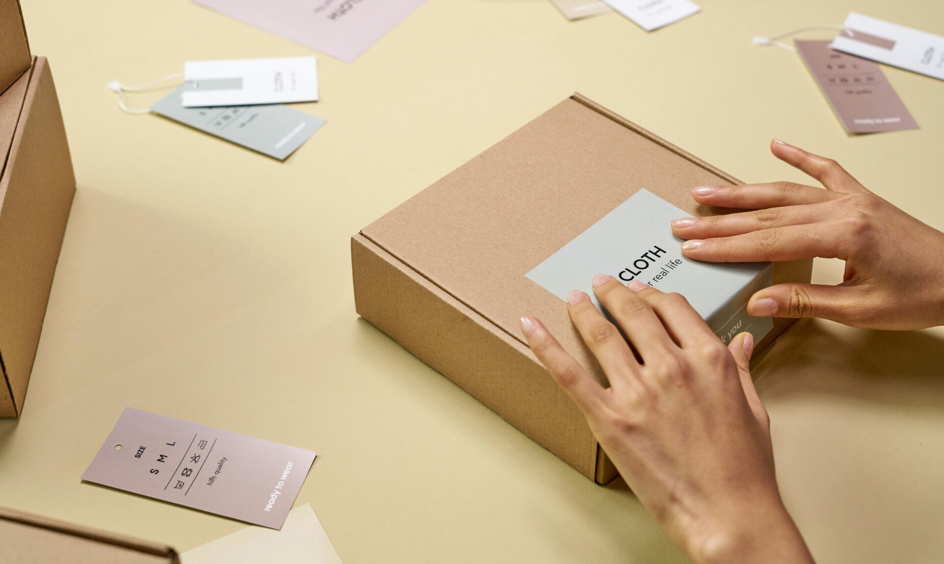 Hands applying a branded label to a small brown e-commerce shipping box, surrounded by tags and product inserts.