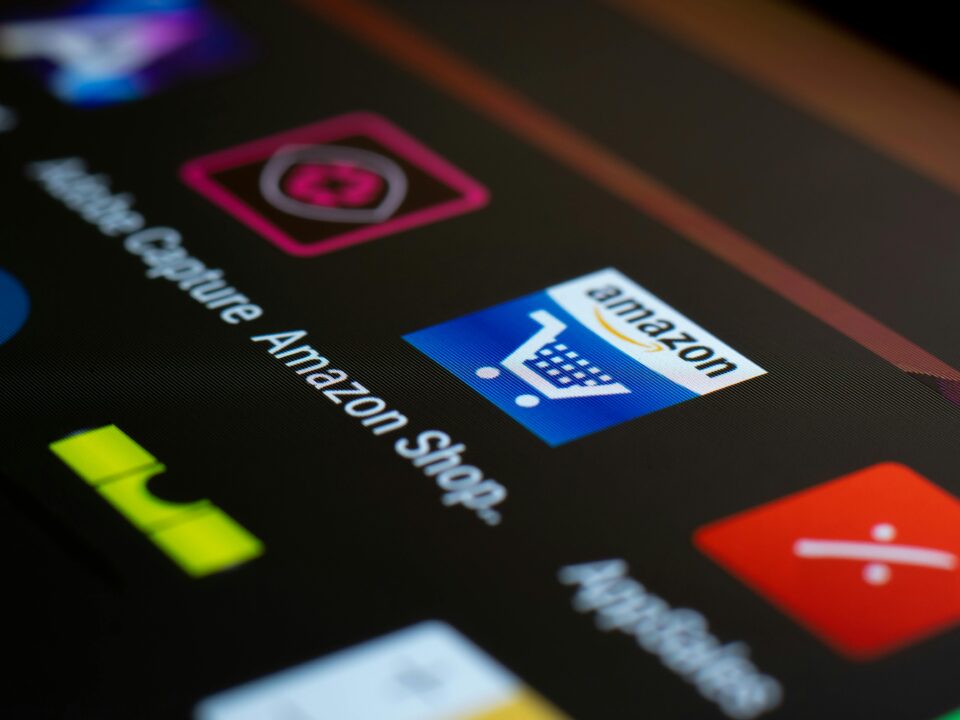 A close-up photograph of a smartphone or tablet screen shows a grid of app icons on a dark background. The central focus is the Amazon Shop app icon, which is blue and features a white shopping cart with the word "amazon" written above it.