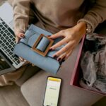 A high-angle shot of a person sitting on a sofa wearing a beige sweater. They are holding a light blue handbag with a large wooden buckle. An open laptop, a smartphone displaying a barcode, and an open pink shipping box with crumpled tissue paper surround them.