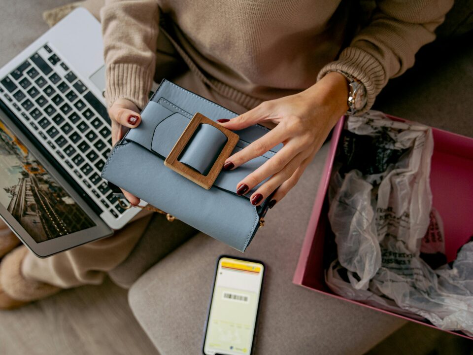 A high-angle shot of a person sitting on a sofa wearing a beige sweater. They are holding a light blue handbag with a large wooden buckle. An open laptop, a smartphone displaying a barcode, and an open pink shipping box with crumpled tissue paper surround them.