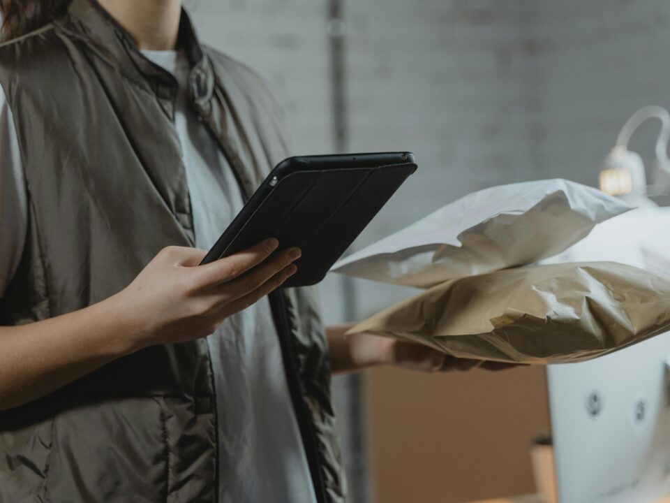 A cropped view of a warehouse or office worker wearing an olive green vest over a white shirt. They are checking a digital tablet held in their left hand while balancing a white and a brown padded mailer in their right hand.