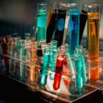 A clear acrylic test-tube rack filled with many test tubes containing brightly colored liquids—reds, blues, greens, oranges, and blacks—illuminated dramatically under laboratory lighting.