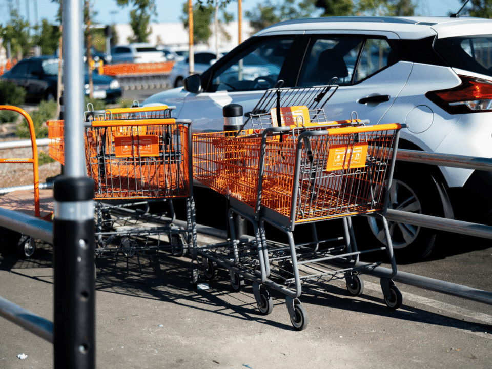 Row of orange shopping carts in a parking area beside a white car on a sunny day, representing retail and e-commerce logistics readiness.