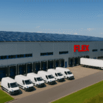 Sustainable FLEX. fulfillment warehouse in Europe with solar panels on the roof, white delivery vans and a truck parked outside under a clear blue sky.