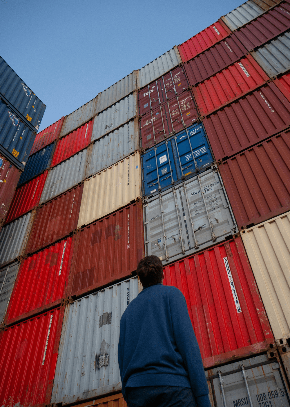 Person observing stacked shipping containers, symbolizing the scale, structure, and global logistics complexity behind enterprise-level supply chain operations.