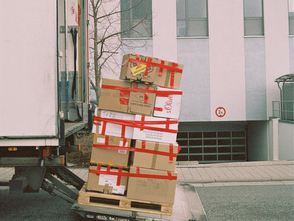 A stack of cardboard boxes secured with red tape is precariously tilted on a loading truck's lift outside a modern building, conveying urgency.
