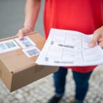 A person in a red shirt holds a package with shipping labels and a printed sheet, standing on a tiled pavement. The scene suggests package delivery.