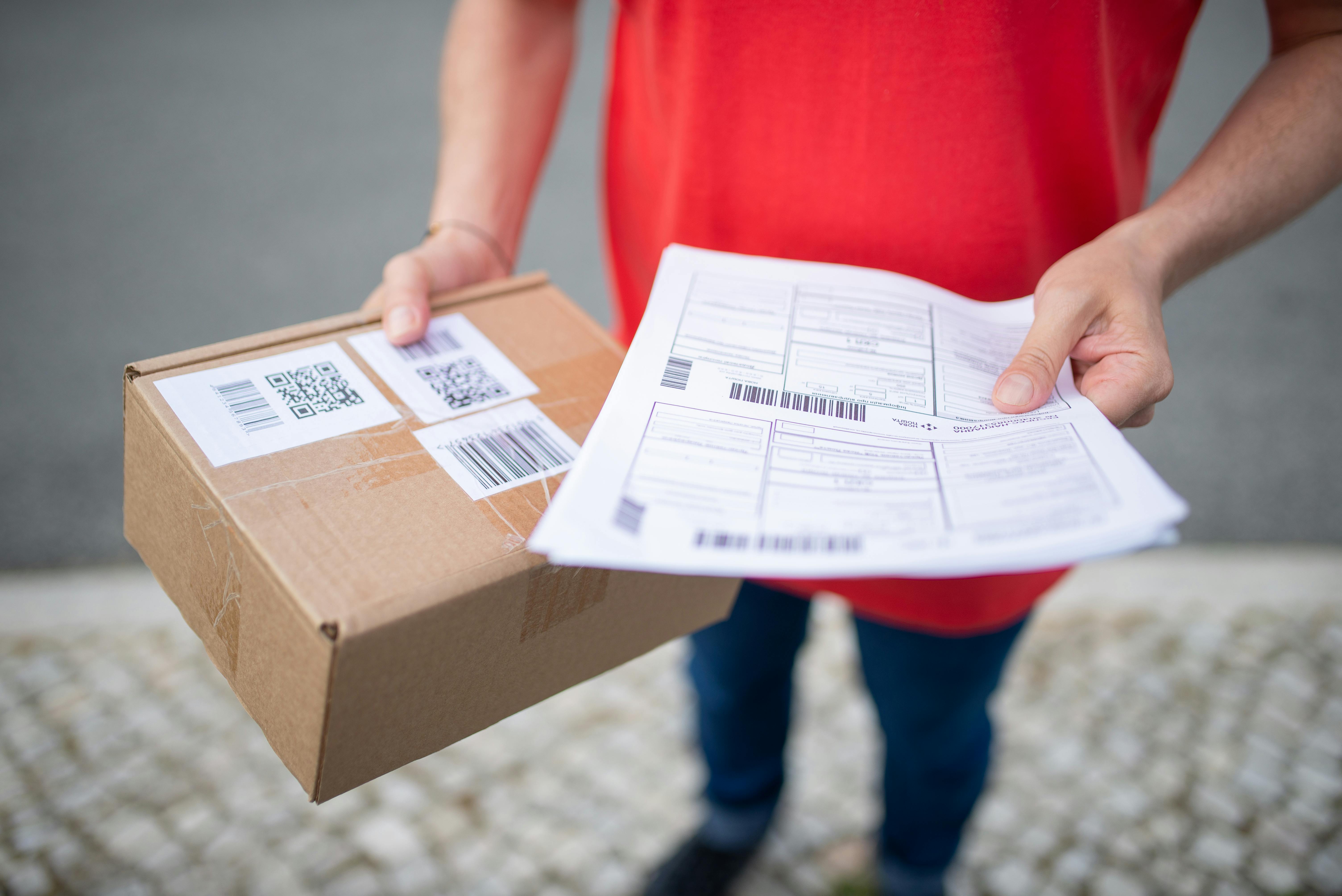 A person in a red shirt holds a package with shipping labels and a printed sheet, standing on a tiled pavement. The scene suggests package delivery.