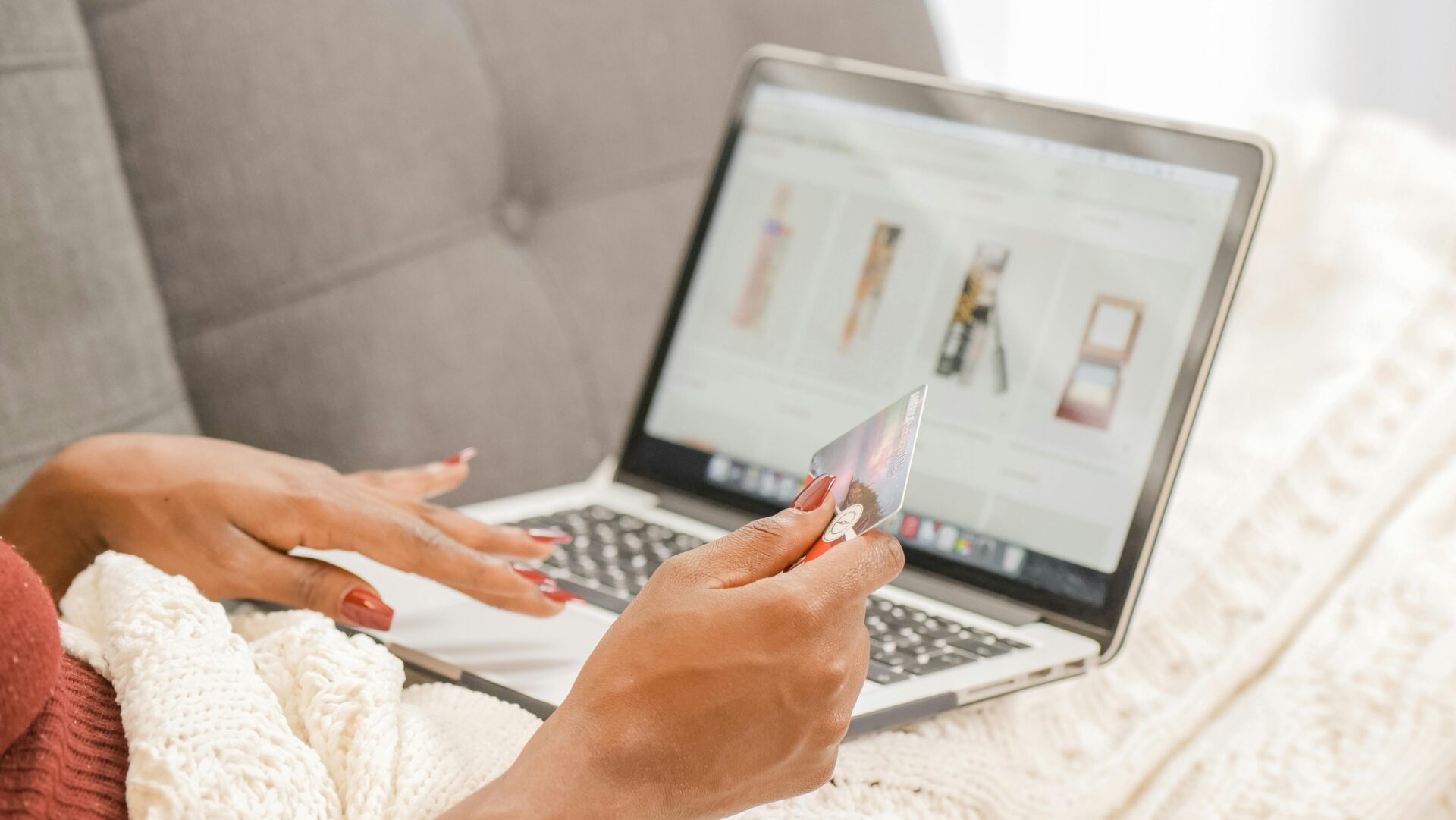 A person with red nails holds a credit card while shopping online on a laptop, displaying beauty products. Cozy setting with knitted blanket and sofa.
