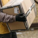 Person in a yellow vest and black gloves carries a large cardboard box marked with shipping labels. The background is a blurred brick wall.