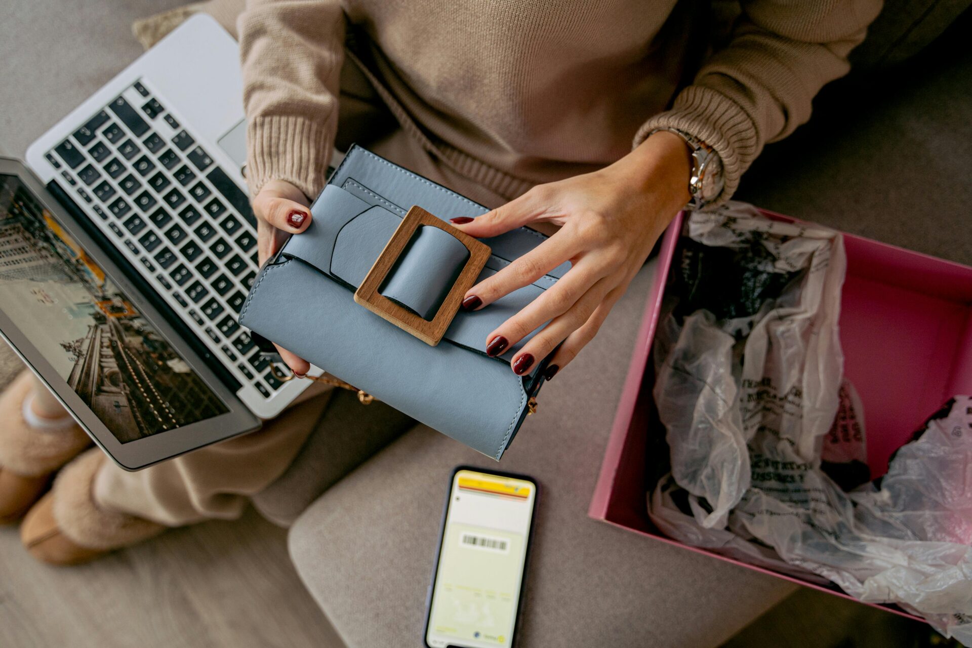 A person examines a blue handbag with a large buckle, sitting on a sofa with a laptop. An open shopping box and a smartphone are nearby. Cozy and engaged atmosphere.