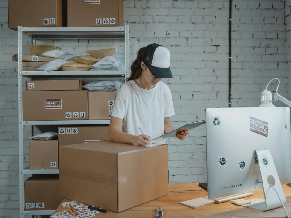 A person in a white shirt and cap checks a clipboard beside a large box in an office. Shelves with packages are in the background, conveying a busy, organized workspace.