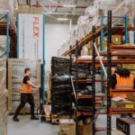 Two workers in orange vests organize packages in a large warehouse with high shelves and stacked boxes. The atmosphere is busy and industrious.