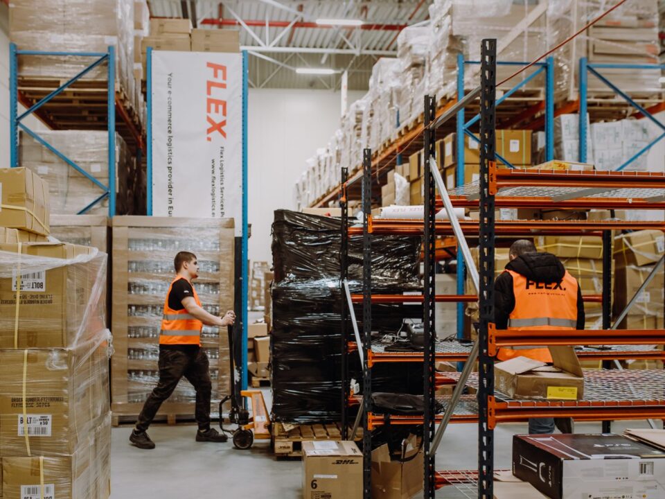 Two workers in orange vests organize packages in a large warehouse with high shelves and stacked boxes. The atmosphere is busy and industrious.