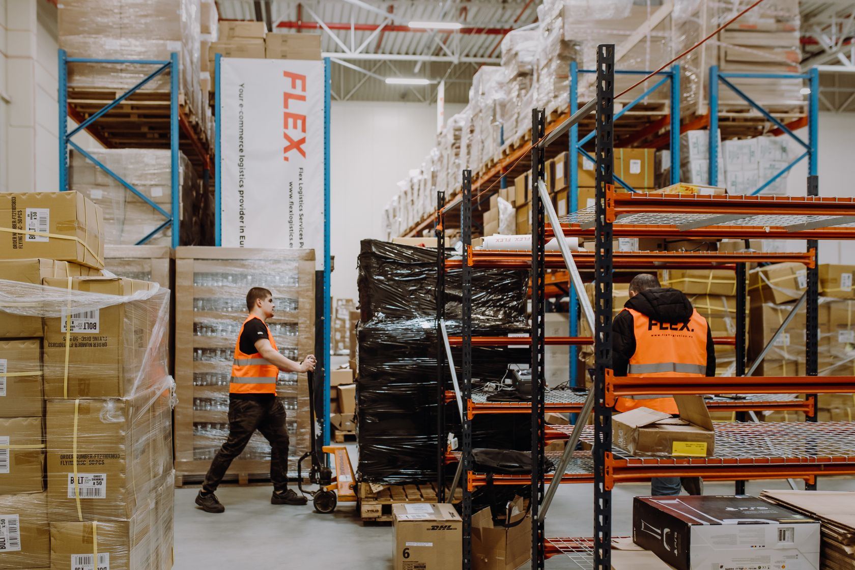 Two workers in orange vests organize packages in a large warehouse with high shelves and stacked boxes. The atmosphere is busy and industrious.
