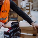 Two workers in orange vests organize boxes and black products in a warehouse. The scene conveys teamwork and efficiency in a busy workspace.