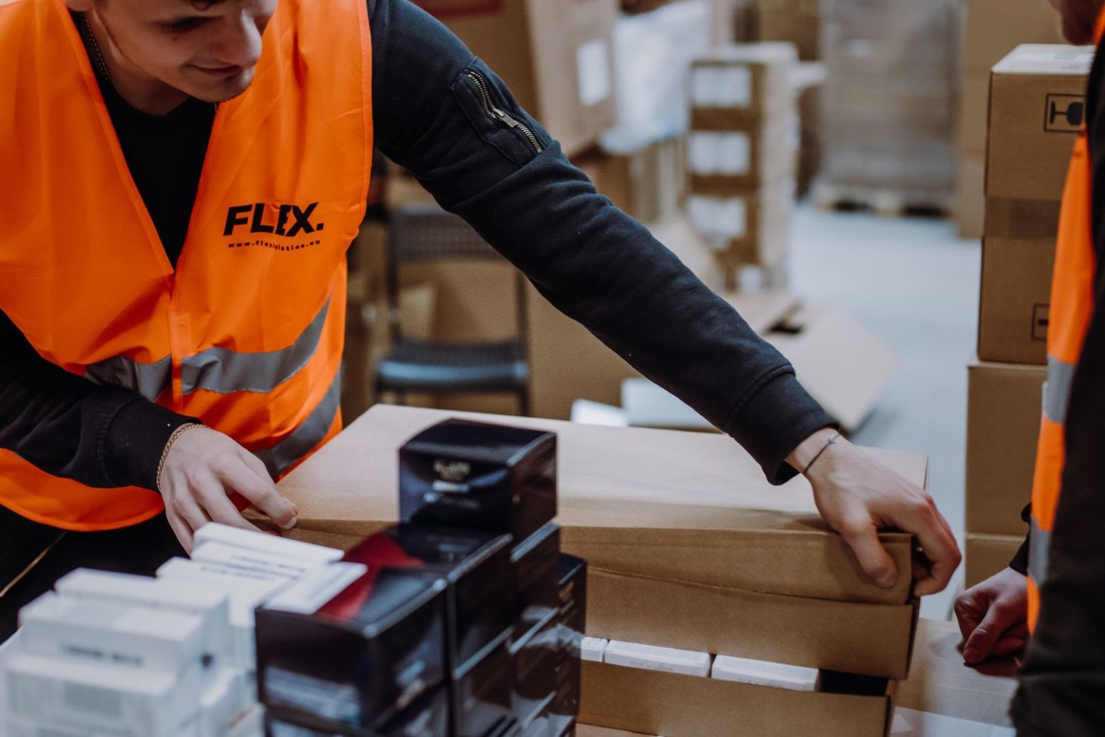 Two workers in orange vests organize boxes and black products in a warehouse. The scene conveys teamwork and efficiency in a busy workspace.