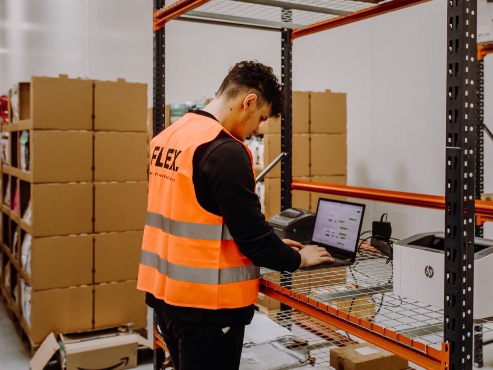 A man in an orange safety vest works on a laptop in a warehouse. He stands near metal shelves filled with cardboard boxes, suggesting an organized setup.