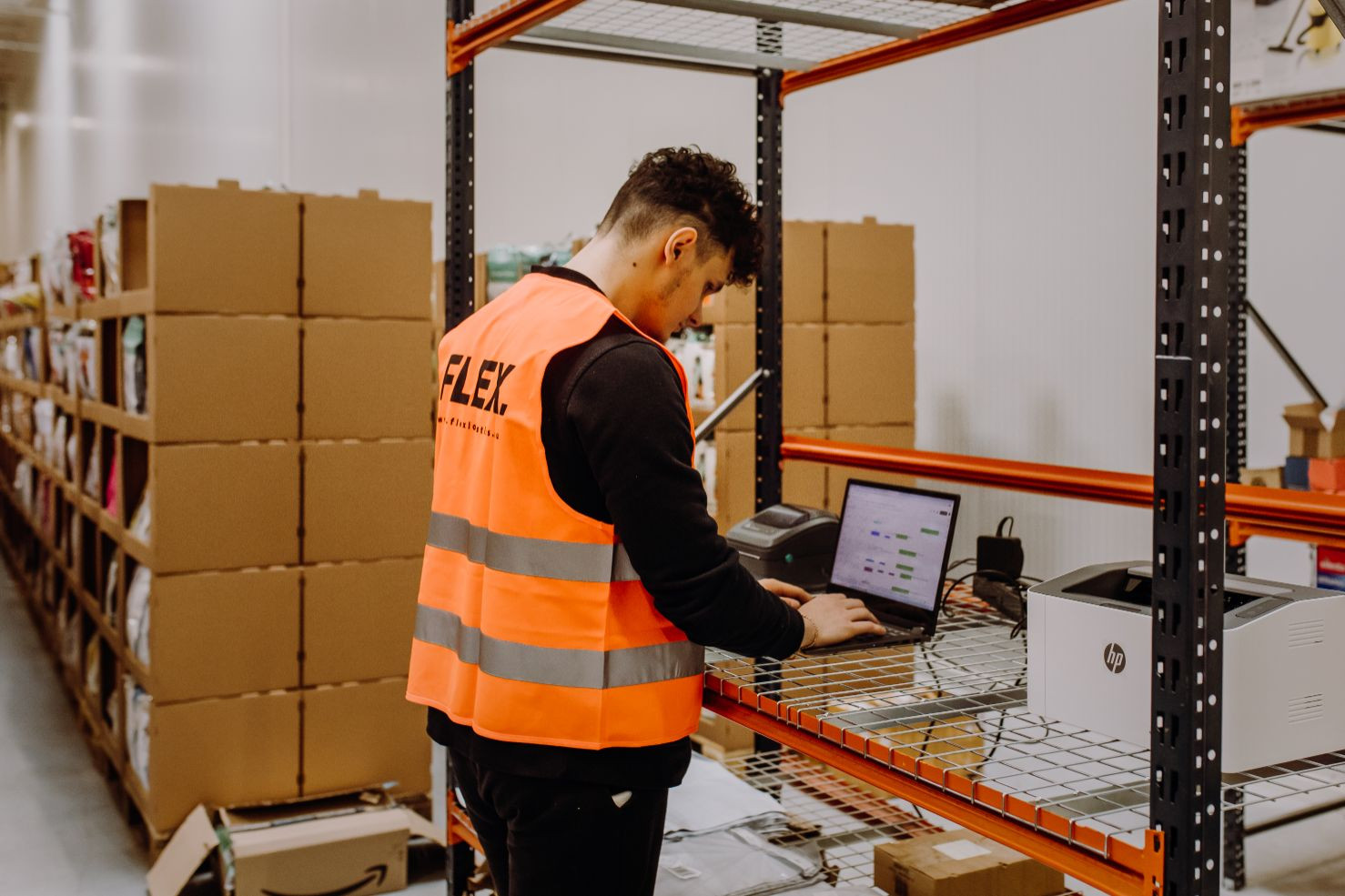 A man in an orange safety vest works on a laptop in a warehouse. He stands near metal shelves filled with cardboard boxes, suggesting an organized setup.