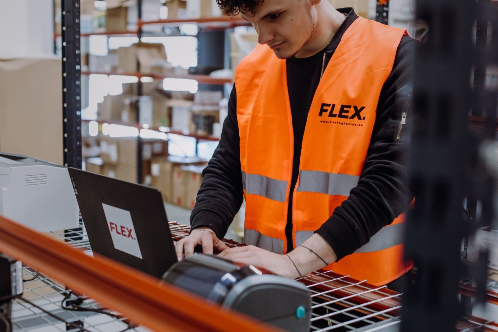 A person in an orange safety vest works on a laptop in a warehouse. Shelves with boxes fill the background, indicating a focused, industrious setting.
