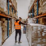 A person wearing an orange safety vest labeled "FLEX" inspects large stacked boxes in a warehouse aisle. The shelves are filled with neatly organized packages.