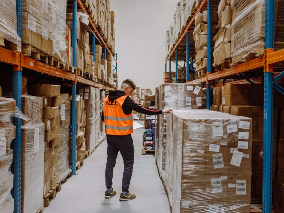 A person wearing an orange safety vest labeled "FLEX" inspects large stacked boxes in a warehouse aisle. The shelves are filled with neatly organized packages.