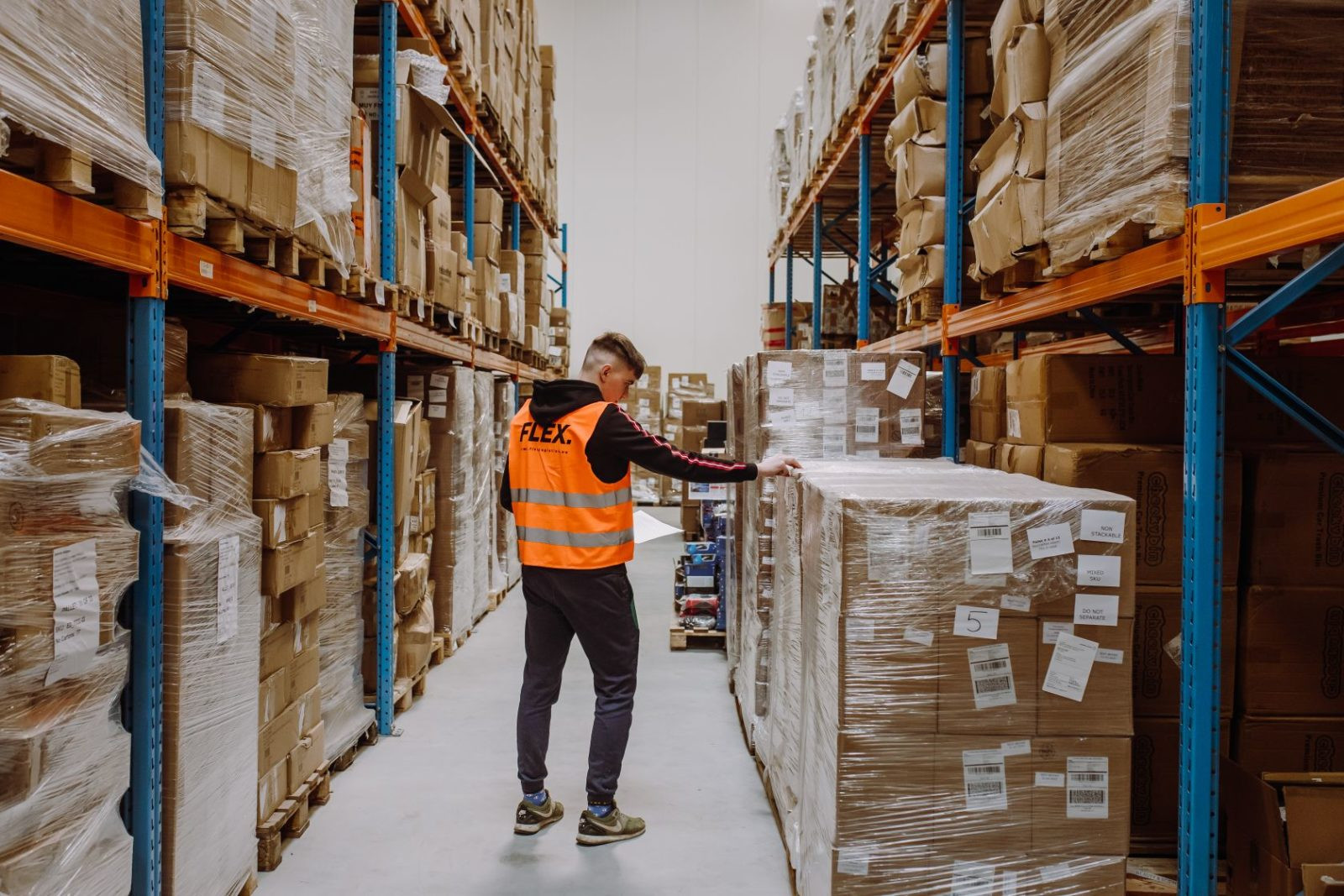 A person wearing an orange safety vest labeled "FLEX" inspects large stacked boxes in a warehouse aisle. The shelves are filled with neatly organized packages.