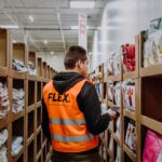 A worker in an orange “FLEX” vest stands in a warehouse aisle surrounded by shelves of packaged garments, holding a phone, focused and attentive.