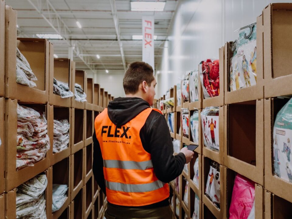 A worker in an orange “FLEX” vest stands in a warehouse aisle surrounded by shelves of packaged garments, holding a phone, focused and attentive.