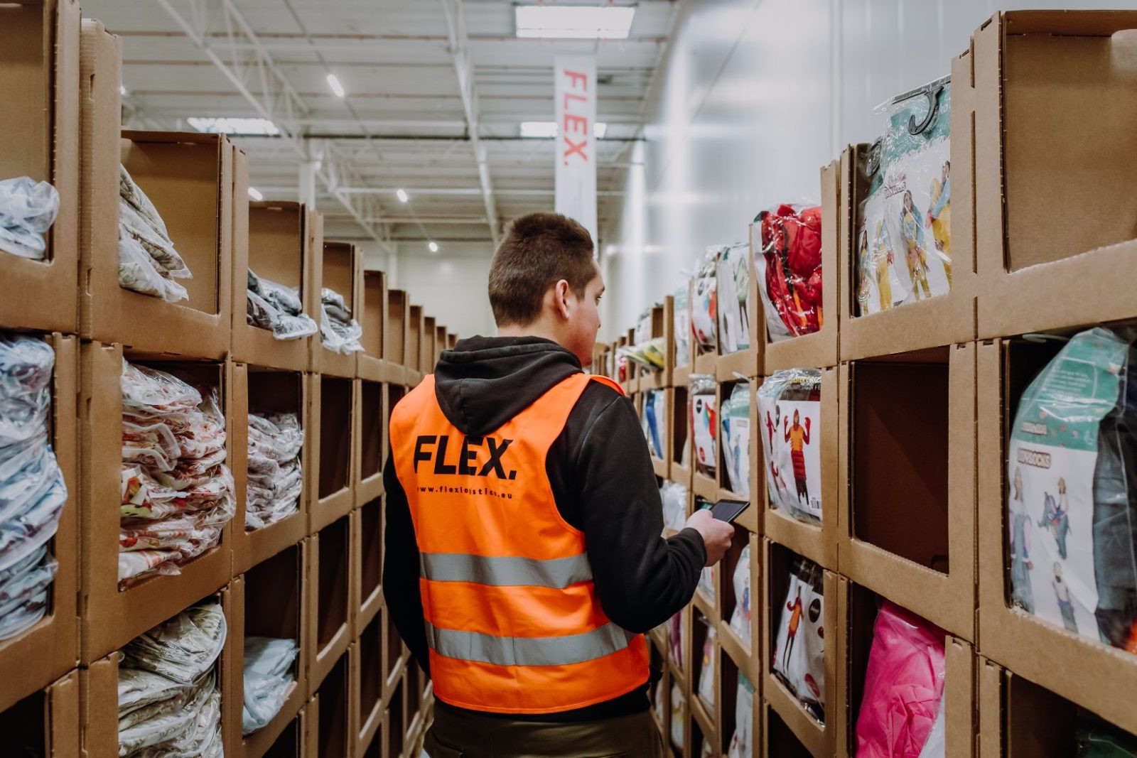 A worker in an orange “FLEX” vest stands in a warehouse aisle surrounded by shelves of packaged garments, holding a phone, focused and attentive.