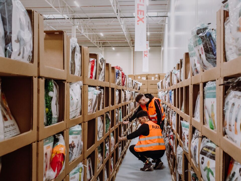 Two workers in orange vests manage inventory between shelves filled with packaged products in a brightly lit warehouse, creating a focused, industrious atmosphere.