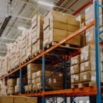 Warehouse interior with tall blue and orange shelves stacked with numerous brown cardboard boxes. Bright lighting and high ceiling create an organized atmosphere.